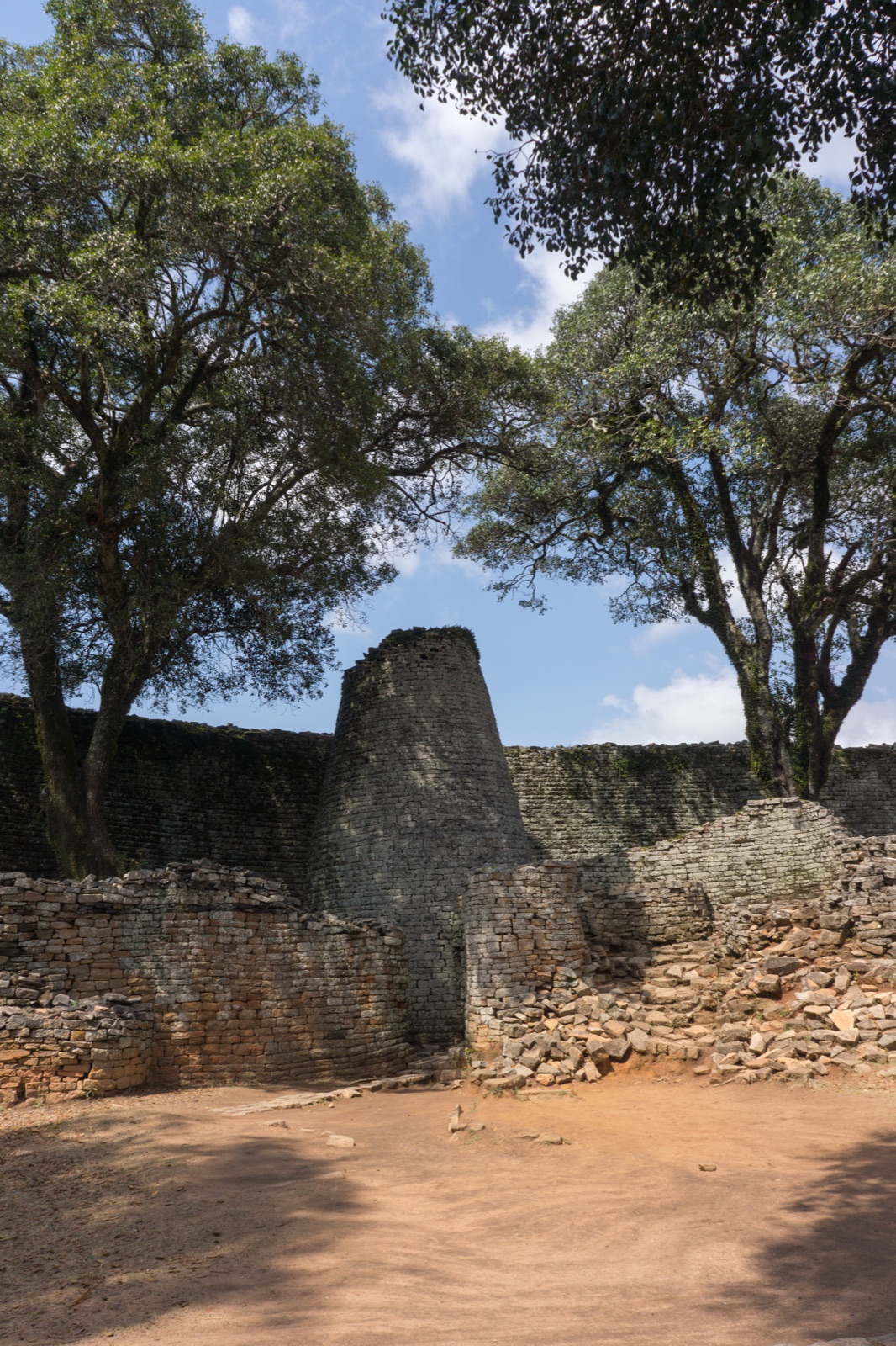 Conical stone tower surrounded by dry stone walls with large trees overhead at Great Zimbabwe archaeological site. Photo: Fanny Schertzer, CC BY 3.0, via Wikimedia Commons.
