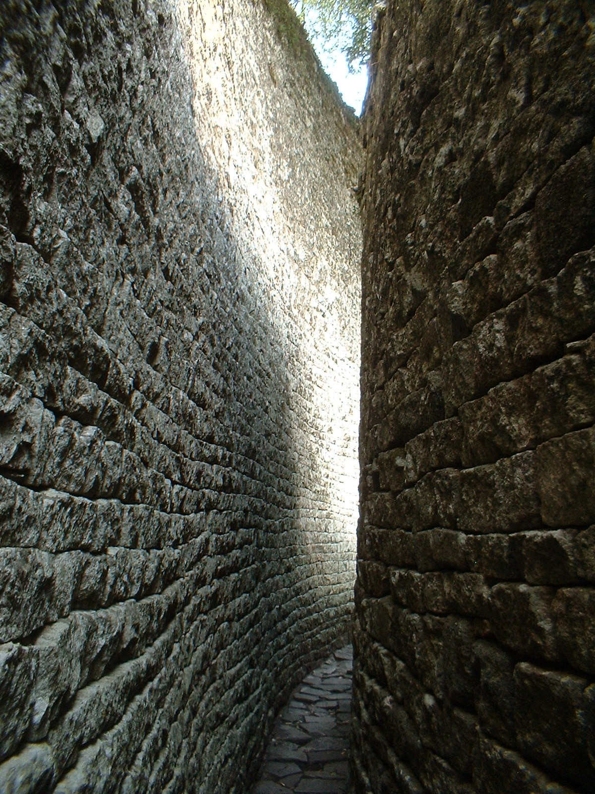 Narrow stone passage between two tall, precisely fitted granite walls at Great Zimbabwe, lit by sky above. Photo: andryn2006, CC BY-SA 2.0, via Wikimedia Commons.