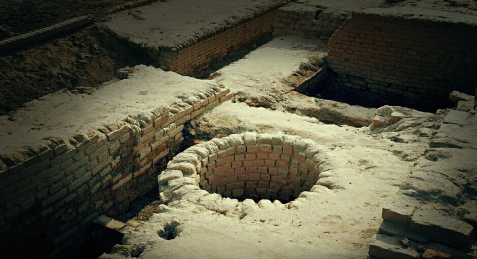 Cylindrical brick-lined well shaft at Mohenjo-daro, its curved terracotta brickwork exposed among excavated walls. Photo: Nikesh chawla, CC BY-SA 4.0, via Wikimedia Commons.