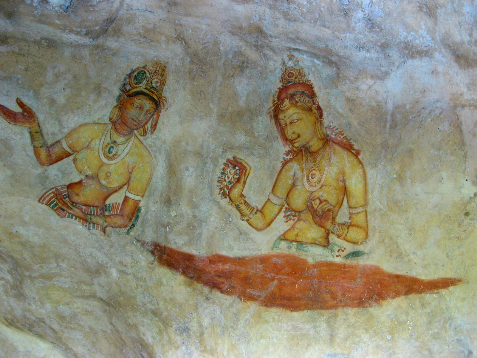 Two ancient frescoes of celestial maidens with ornate headdresses and jewelry painted on a weathered rock face at Sigiriya. Photo: Michael Gunther, CC BY-SA 4.0, via Wikimedia Commons.